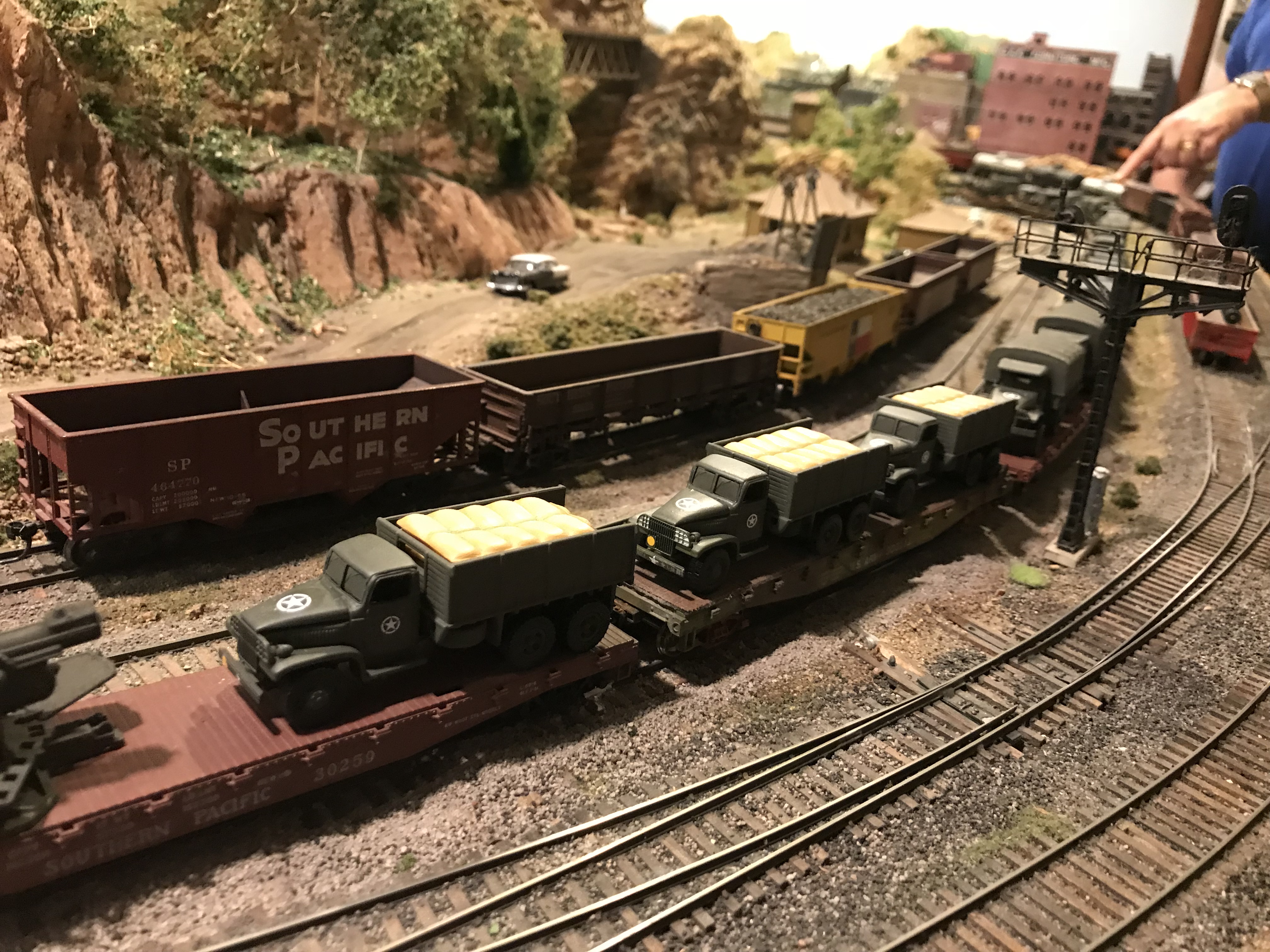 HO scale 3D printed olive drab military trucks with canvas cargo loads on flatcars, passing a Southern Pacific gondola