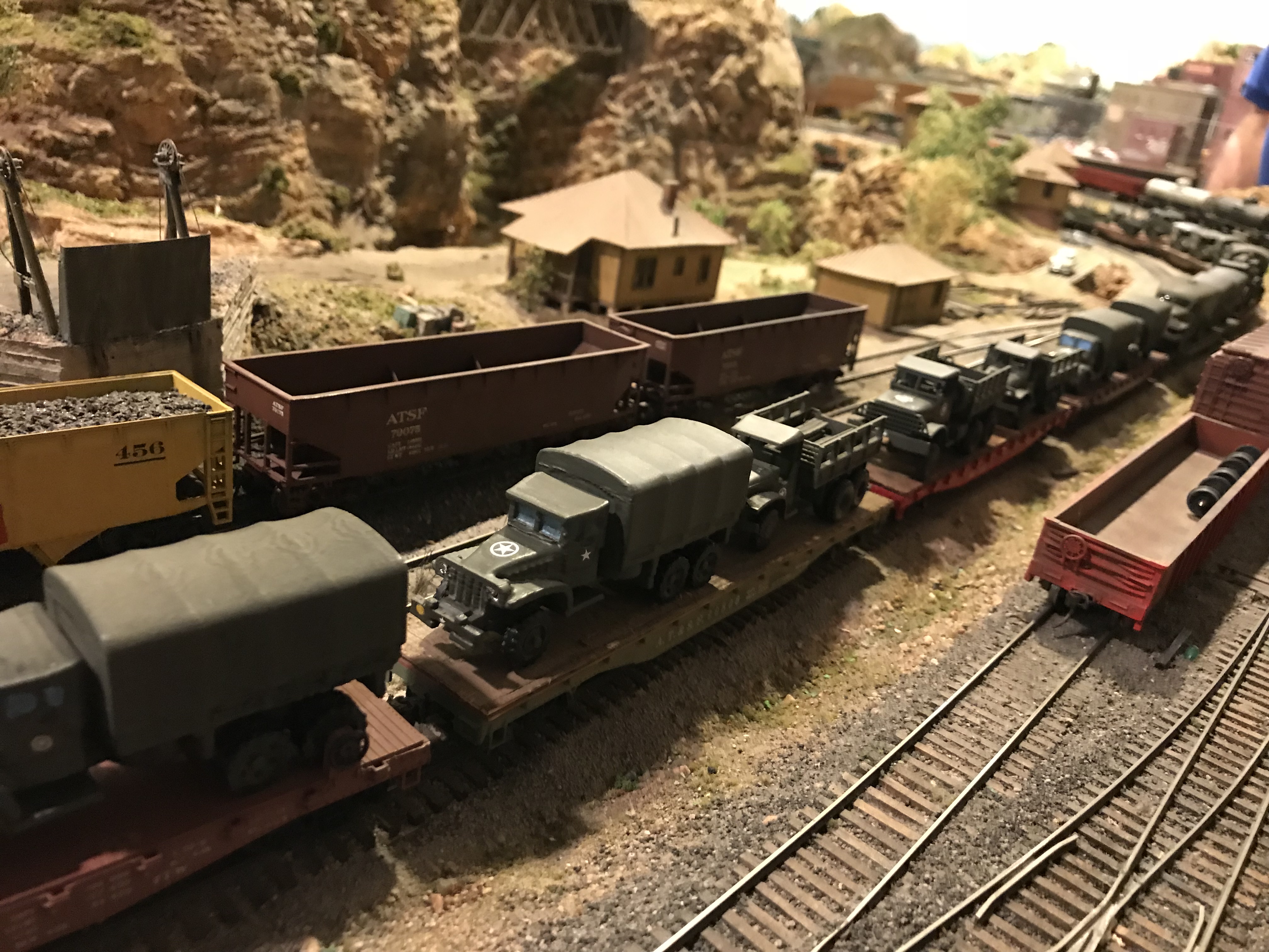 Close up view of the military train's 3D printed trucks on flatcars, passing an ATSF gondola and yellow 456 gondola near a layout structure