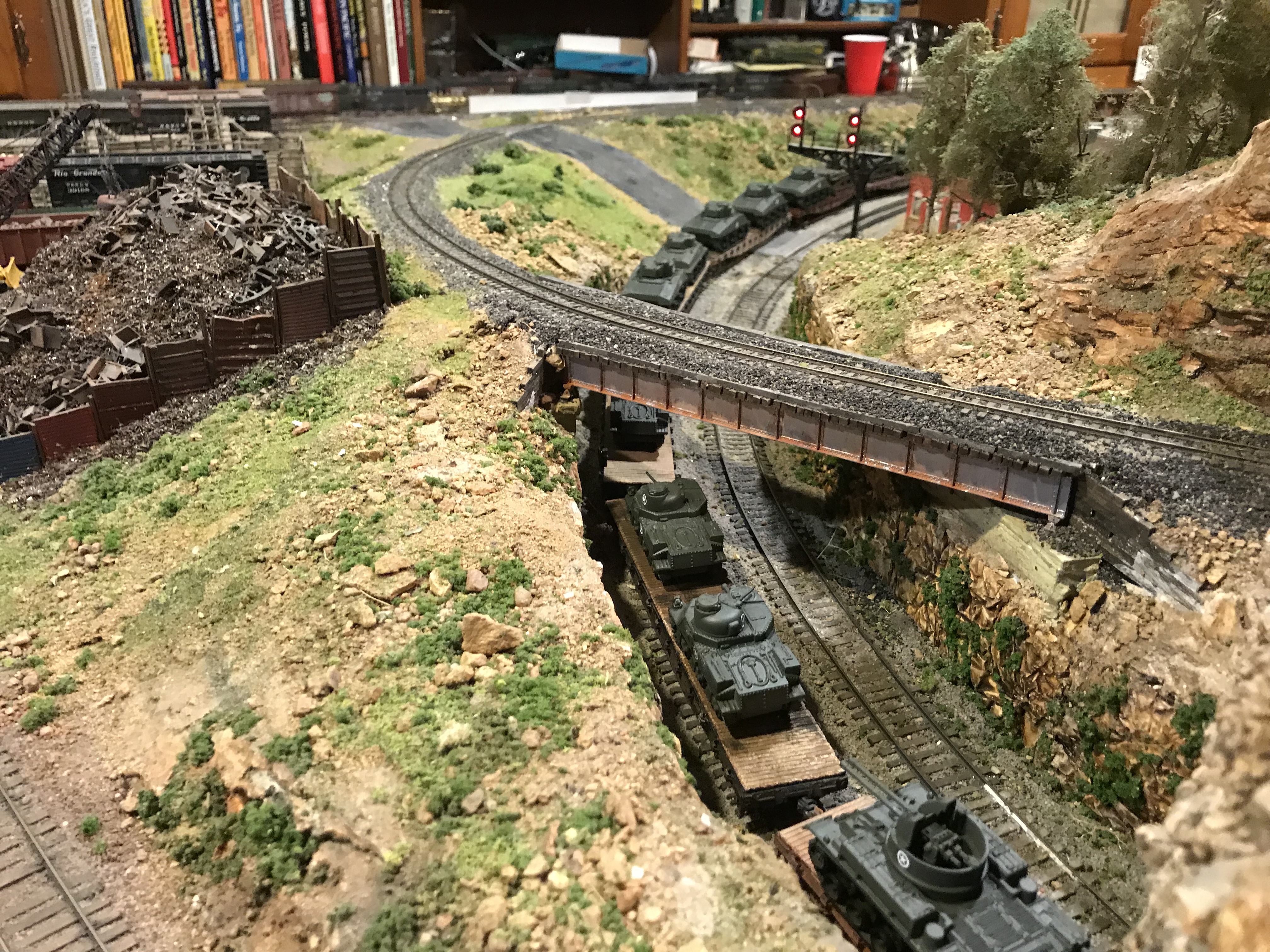 Elevated view of a 3D printed Sherman tank train moving on a curved track, passing under a bridge near a large scrap metal pile