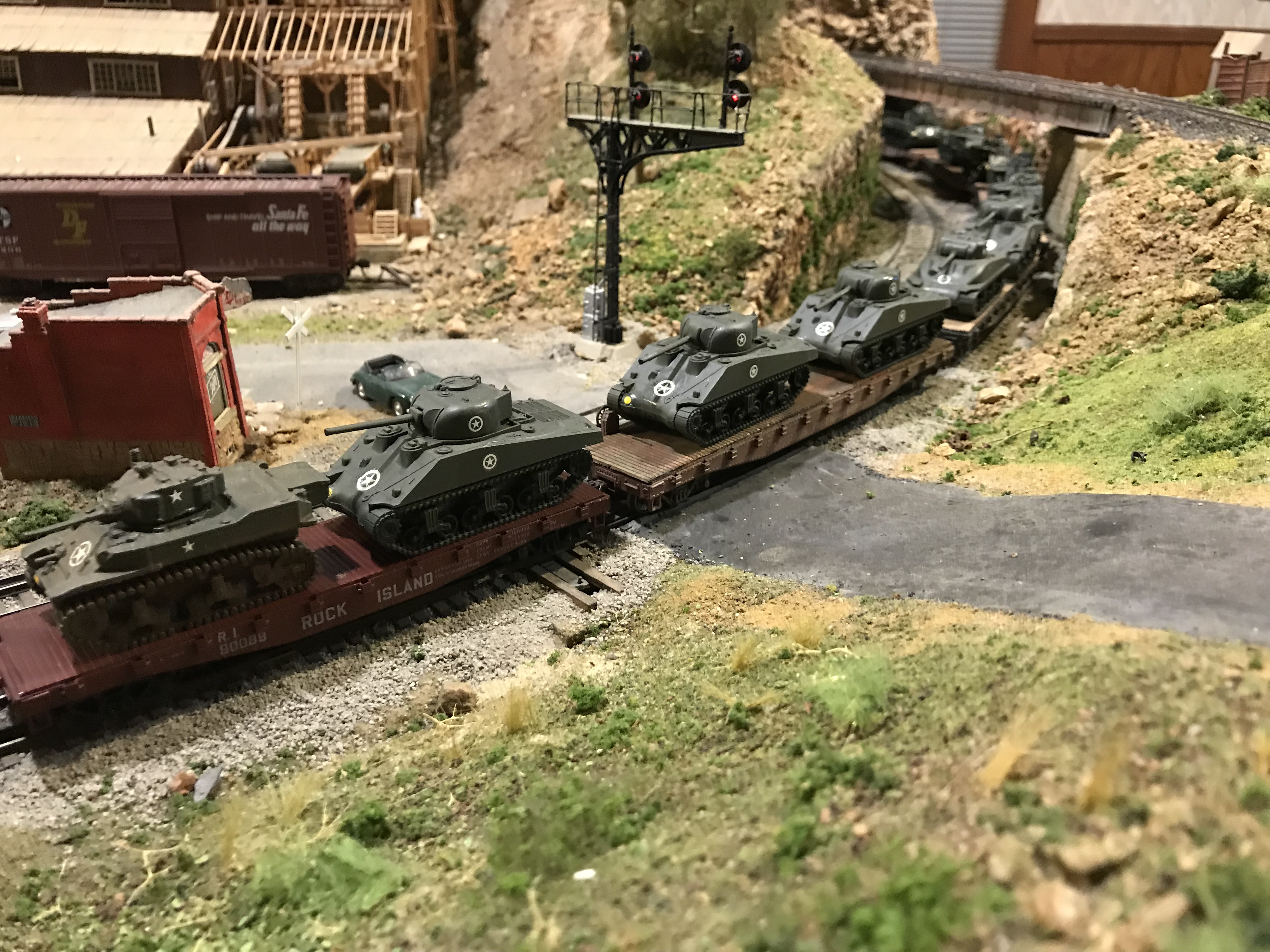 The model military train with Sherman tanks crossing a layout road, headed towards a bridge with a large mill structure in the background