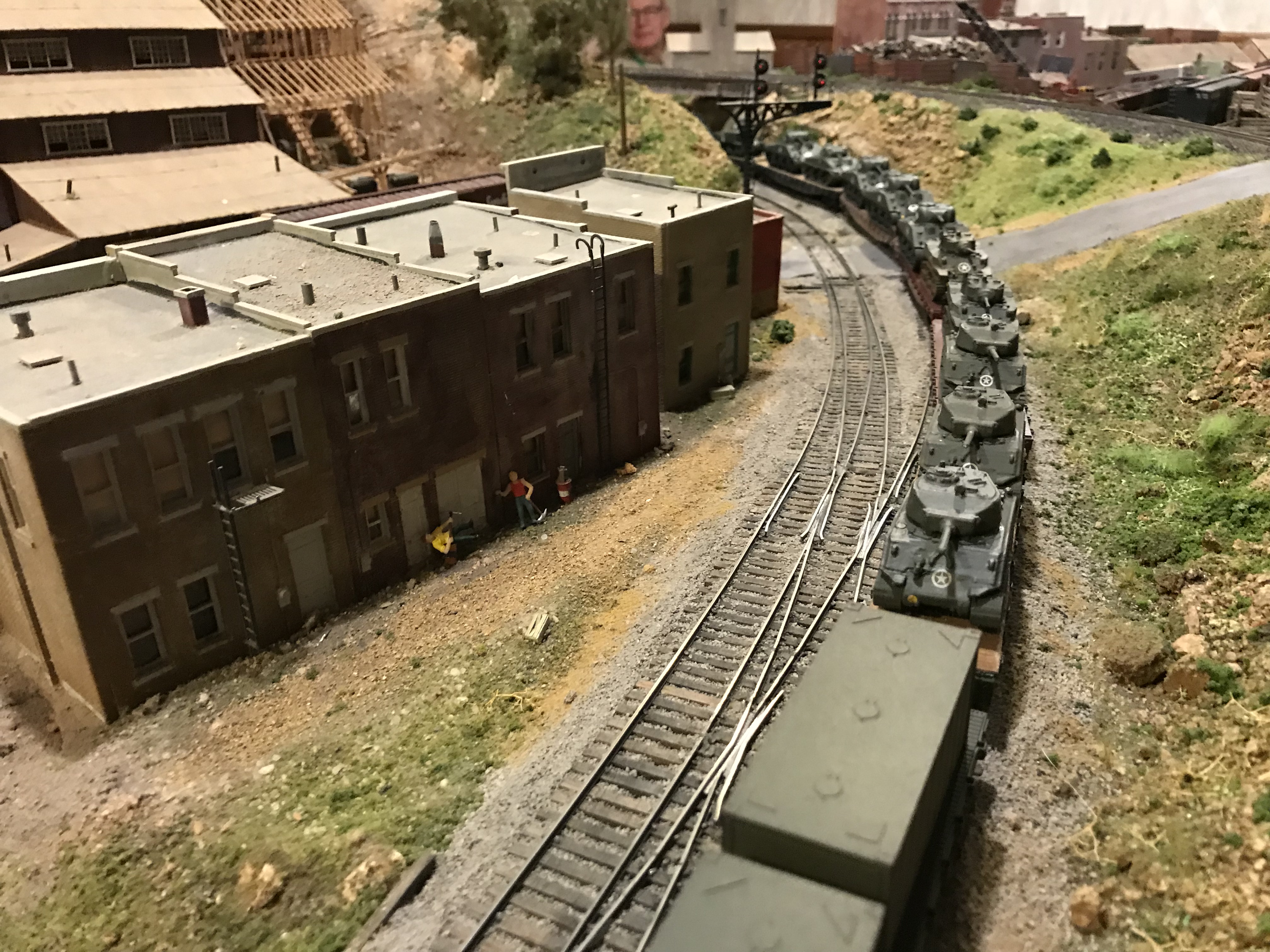High angle perspective looking down a curved line of flatcars loaded with 3D printed Sherman tanks, passing miniature brick town buildings